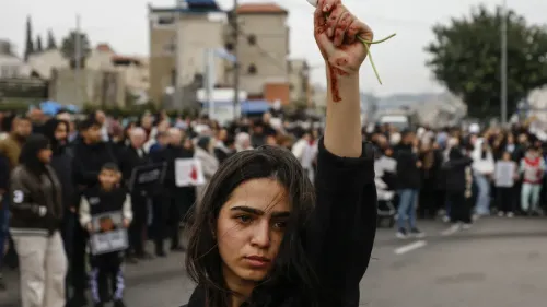 امرأة فلسطينية ترفع يدها عالياً وهي تحمل غصنًا أخضر، بينما تظهر على وجهها علامات الدم، وسط حشد من المتظاهرين في الخلفية.