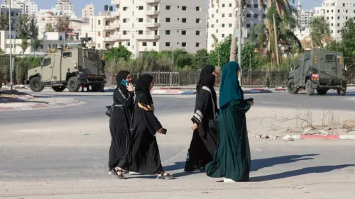 نساء يرتدين الملابس التقليدية يتجولن في منطقة حضرية، مع وجود مركبة عسكرية في الخلفية، تعكس الأوضاع الأمنية في الضفة الغربية.