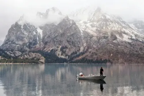 شخص يقف في قارب صغير على بحيرة هادئة، محاطًا بجبال غراند تيتون المغطاة بالثلوج والسحب.