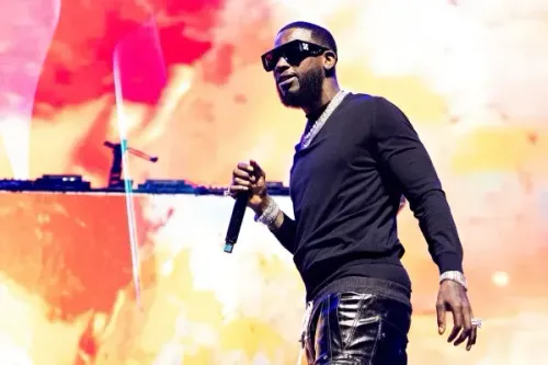 مغني الراب غوتشي ماني يؤدي على المسرح، يرتدي نظارات شمسية وقميصًا أسود، مع خلفية ملونة تعكس أجواء الحفل.