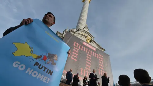 رجل يحمل لافتة زرقاء مكتوب عليها "PUTIN GO HOME!" خلال فعالية في كييف، مع نصب تذكاري بارز في الخلفية.