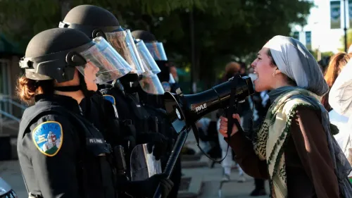 امرأة ترتدي حجابًا وتستخدم مكبر صوت للاحتجاج، تواجه صفًا من رجال الشرطة المدججين بالأسلحة، في مشهد يعكس التوتر بين المتظاهرين والسلطات.