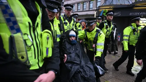 احتجاجات في لندن حيث تقوم الشرطة باعتقال شخص يرتدي غطاءً أسود، وسط تواجد مكثف للشرطة. تعكس الصورة التوترات حول القوانين الجديدة المتعلقة بالاحتجاجات.
