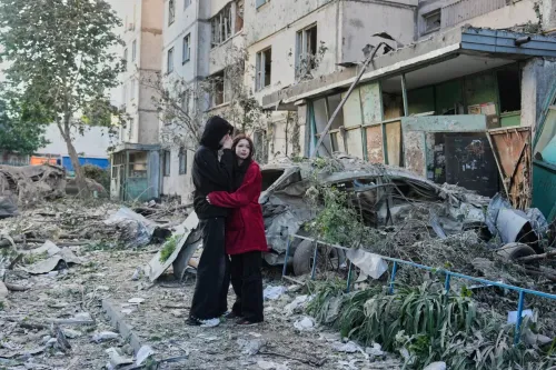زوجان يحتضنان بعضهما في منطقة مدمرة، حيث تظهر آثار الهجوم الروسي على المباني والسيارات المحطمة في الخلفية.