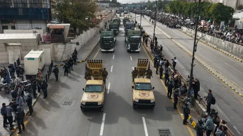 عرض عسكري في الشارع، حيث تسير شاحنات عسكرية ومركبات مدرعة، بينما يتجمع الناس على جوانب الطريق.