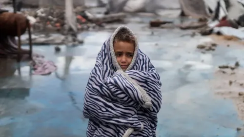 طفل فلسطيني wrapped in a striped blanket، يبدو حزينًا ومرتبكًا وسط أنقاض المنازل المدمرة في غزة، مع تدهور الأوضاع الإنسانية.