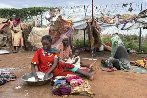 نساء وأطفال في مخيم للنازحين بجنوب السودان، حيث تقوم امرأة بغسل الملابس بينما يجلس الآخرون في الخلفية وسط ظروف صعبة.
