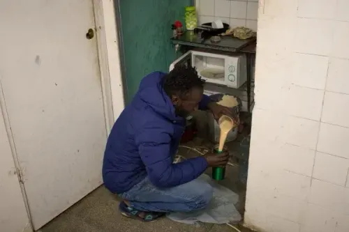 رجل يرتدي سترة زرقاء يجلس على الأرض في مطبخ مهجور، يقوم بصب مكونات في وعاء، مما يعكس ظروف المعيشة الصعبة للمهاجرين في بادالونا.