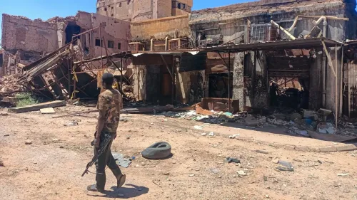 رجل مسلح يسير في منطقة مدمرة، تظهر خلفه مبانٍ متهدمة وأرض مليئة بالحطام، تعكس آثار النزاع في السودان.