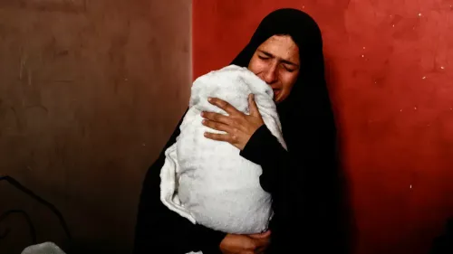 امرأة ترتدي الحجاب تحمل طفلاً ملفوفًا ببطانية، تعبر عن حزن عميق في غرفة ذات جدران حمراء، تعكس معاناة عائلتها في غزة.