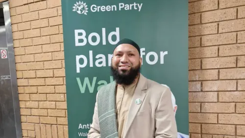 مؤذن علي، نائب رئيس حزب الخضر، مبتسم أمام لافتة الحزب، يعبر عن قلقه من التهديدات بالقتل بعد ادعاءات كاذبة ضده.