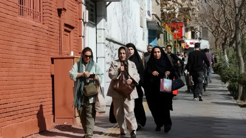 نساء إيرانيات يرتدين الحجاب ويمشين في شارع مزدحم، مع وجود أشجار ومباني في الخلفية، تعكس الحياة اليومية في طهران.