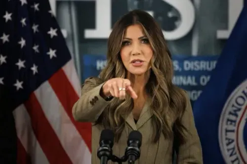 امرأة تتحدث في مؤتمر صحفي، تشير بإصبعها، مع أعلام خلفها، تعكس تعزيز إنفاذ قوانين الهجرة في إدارة ترامب.