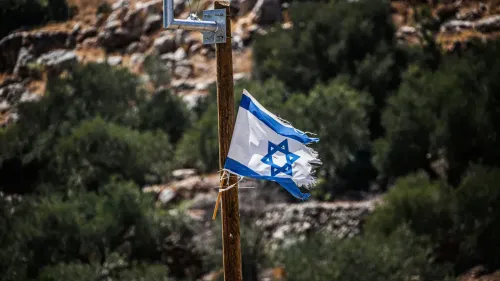 علم إسرائيلي ممزق يرفرف على عمود خشبي في منطقة جبلية، محاط بأشجار خضراء، في سياق التوترات حول الضفة الغربية.