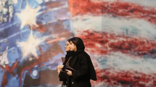 امرأتان ترتديان الحجاب تسيران معًا أمام جدارية تحمل ألوان العلم الأمريكي، مما يعكس التوترات السياسية بين إيران والولايات المتحدة.