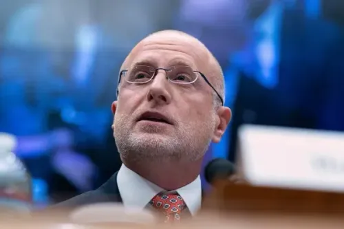 رجل ذو لحية وشعر رمادي، يرتدي نظارات وقميصًا أبيض، يتحدث خلال جلسة استماع رسمية، مع خلفية تضم شاشات عرض.