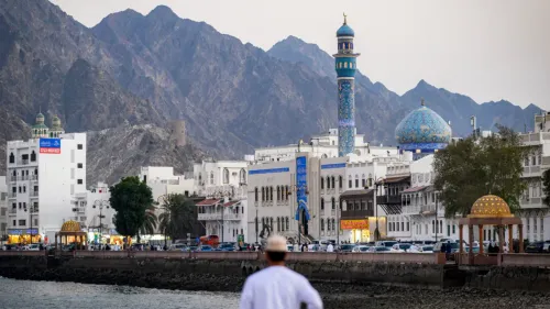 تظهر الصورة مشهداً لمدينة مسقط، حيث تتداخل المباني الحديثة مع الجبال، وتبرز المآذن الملونة، مما يعكس التراث الثقافي لعمان.
