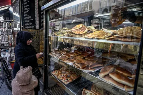 امرأة ترتدي الحجاب تتصفح هاتفها أمام واجهة عرض لمخبز، حيث تظهر أنواع مختلفة من الخبز المسطح والمعجنات.