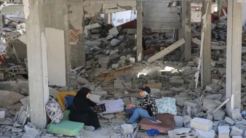 امرأتان تجلسان على وسائد بين أنقاض مستشفى مدمر، مع تواجد الحطام حولهما، مما يعكس آثار الدمار والخراب.