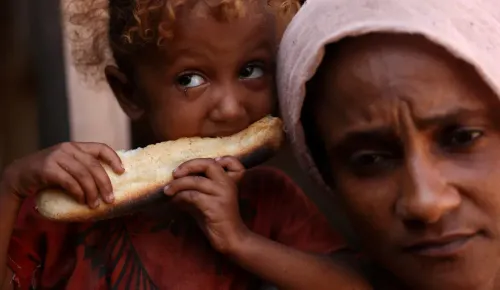 طفل يمسك بقطعة خبز ويأكل منها، بينما تظهر والدته بملامح قلق. تعكس الصورة معاناة الأسر في اليمن بسبب الأوضاع الإنسانية الصعبة.