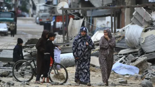 نساء وأطفال يسيرون في منطقة مدمرة في غزة، مع بقايا المنازل المهدمة حولهم، مما يعكس آثار الصراع المستمر.