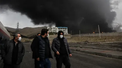 دخان أسود كثيف يتصاعد من موقع انفجار في إيران، مع وجود مجموعة من الأشخاص يرتدون أقنعة واقية في المقدمة، مما يعكس تصاعد التوترات في المنطقة.