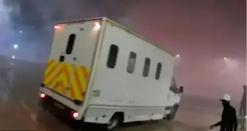 سيارة نقل محصنة تخرج من موقع مظلم، مع وجود ضباب كثيف حولها، بينما يتواجد أشخاص يرتدون ملابس داكنة في الخلفية.