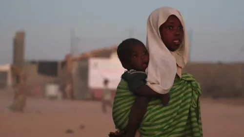 امرأة تحمل طفلاً على ظهرها، ترتدي حجابًا، في خلفية تعكس ظروف النزوح في السودان بعد تصاعد العنف.