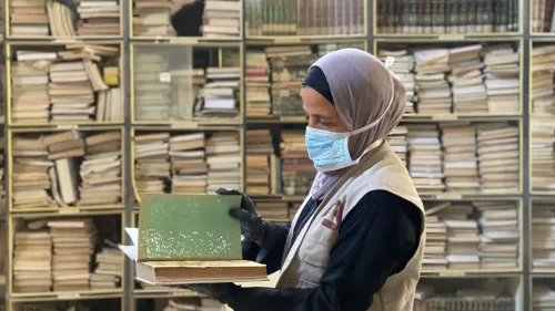 امرأة فلسطينية ترتدي كمامة، تحمل كتابًا متضررًا في مكتبة المسجد العمري الكبير بغزة، وسط رفوف مليئة بالكتب. جهود إنقاذ التراث الثقافي مستمرة رغم التحديات.