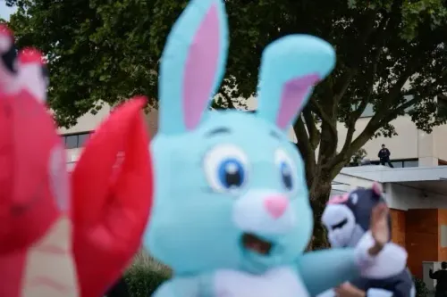 شخصيات ملونة من كرتون، تشمل أرنبًا أزرق كبيرًا، تظهر في حدث عام مع وجود شخص يرتدي زيًا رسميًا في الخلفية.