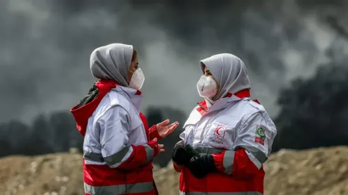 امرأتان ترتديان ملابس طبية وقائية تتحدثان في موقع حرائق النفط بطهران، مع تصاعد الدخان الكثيف في الخلفية، مما يعكس آثار الهجمات على البيئة وصحة السكان.