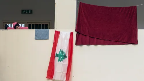 طفلة لبنانية ترتدي سترة وردية، تستند إلى حائط، بجوار علم لبنان ومناشف معلقة، تعكس الظروف الإنسانية الصعبة في لبنان.
