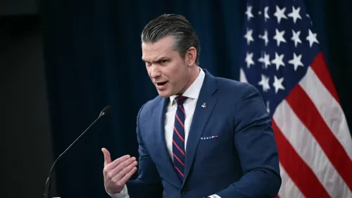 رجل يتحدث بحماس أمام ميكروفون، يرتدي بدلة زرقاء، وخلفه علم الولايات المتحدة، يعبر عن آراء حول الحرب على إيران وتأثيرها الديني.