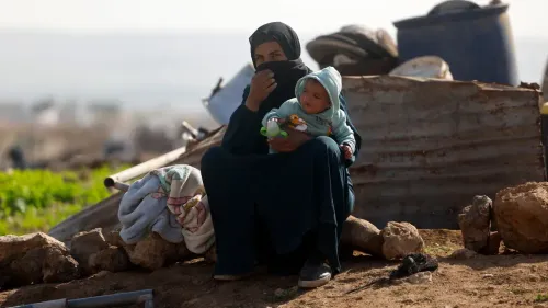 امرأة فلسطينية ترتدي الحجاب، تحمل طفلًا في حضنها، تجلس بجوار أكوام من الأقمشة والأدوات، تعكس معاناة الحياة اليومية في الضفة الغربية.