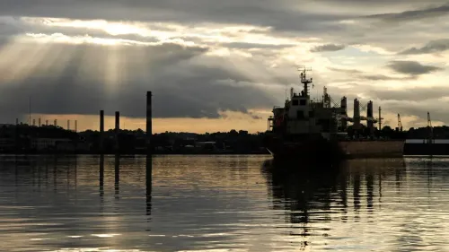 سفينة شحن تحمل النفط الفنزويلي تبحر في مياه هادئة تحت سماء غائمة، مع ظهور أعمدة دخان في الخلفية، تشير إلى استئناف صادرات النفط.