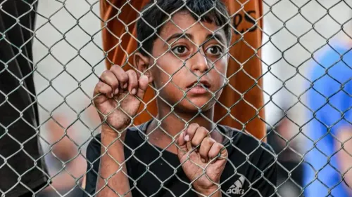 طفل فلسطيني ينظر من خلال سياج معدني، معبرًا عن مشاعر الخوف والحرمان في ظل ظروف الاحتلال.