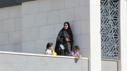 امرأة فلسطينية ترتدي عباءة تقف على شرفة منزل محاصر في قصرة مع طفلين، تعكس معاناة الحصار ونقص الغذاء في الضفة الغربية.
