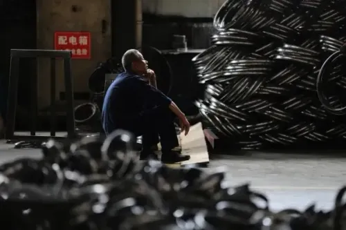 رجل يجلس في مصنع، محاط بأجزاء معدنية، يعكس التحديات الاقتصادية التي تواجه الصين مع تزايد الضغوط التجارية والركود في قطاع العقارات.