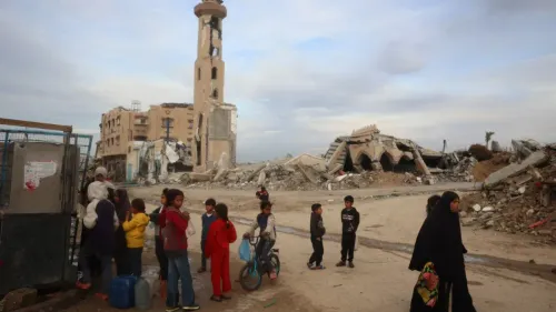 أطفال ونساء يقفون بجوار أنقاض مبانٍ مدمرة في غزة، مع بقايا مئذنة مسجد شاهدة على الدمار الناتج عن النزاع.