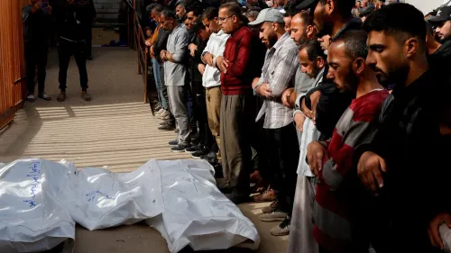 تجمع حشد من الرجال في جنازة، يحيطون بجثتين مغطاتين بأكياس بيضاء، تعبيرات الحزن واضحة على وجوههم.