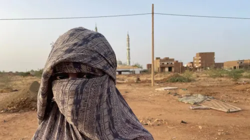 امرأة ترتدي غطاء رأس تقليدي، تقف في منطقة شبه صحراوية، مع مبانٍ خلفها وأحد المساجد في الأفق، تعكس معاناة سكان الصالحة في السودان.