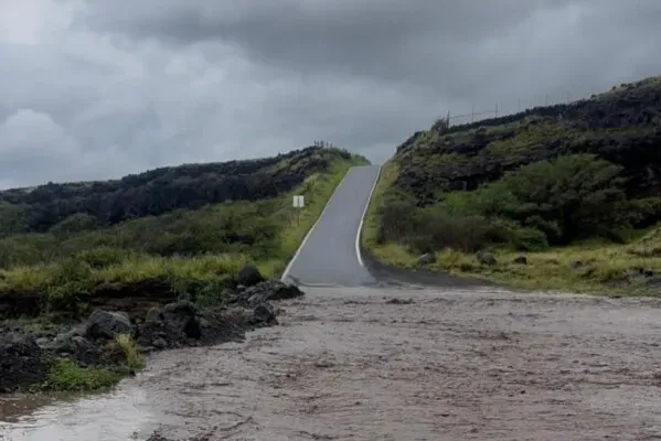 طريق مغمور بالمياه بسبب الفيضانات في هاواي، مع سماء غائمة وأرض محيطة خضراء، مما يعكس تأثير الطقس القاسي على المنطقة.