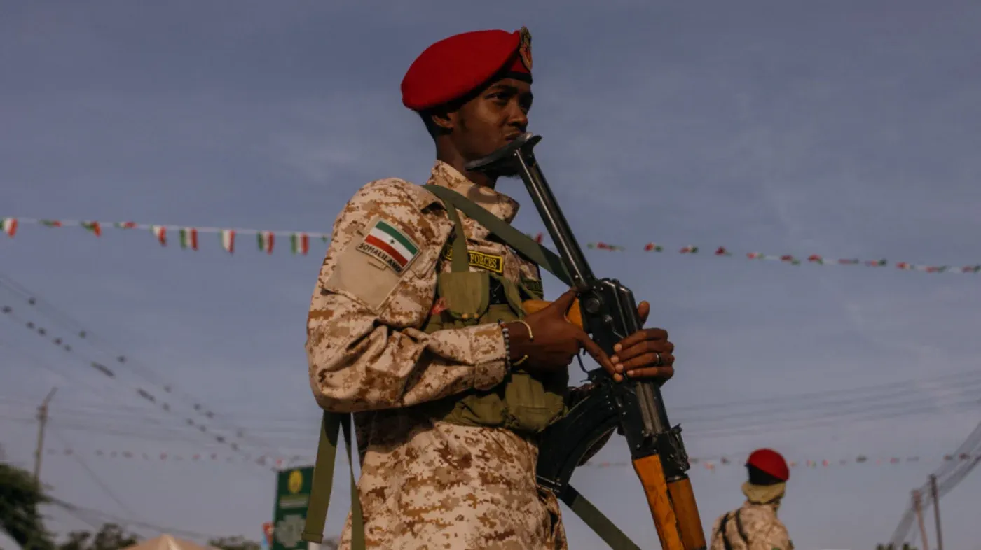 جندي من صومالي لاند يرتدي زيًا عسكريًا ويحمل سلاحًا، مع خلفية تظهر الأعلام الملونة، تعبيرًا عن الاستعدادات للاحتفال بالاستقلال.