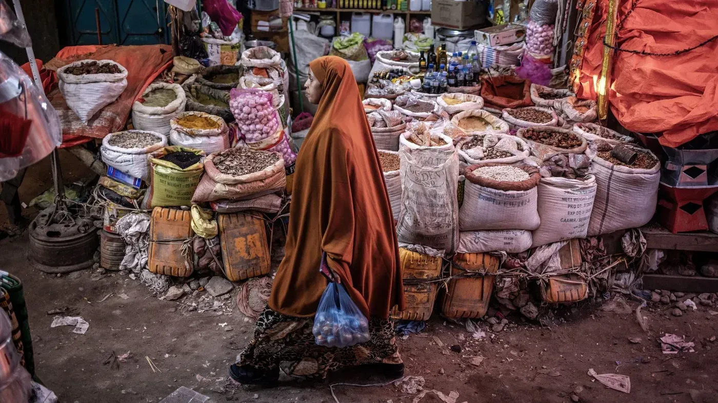 امرأة ترتدي عباءة برتقالية تسير في سوق هرجيسا، محاطة بأكياس من التوابل والمواد الغذائية، تعكس الحياة اليومية في أرض الصومال.