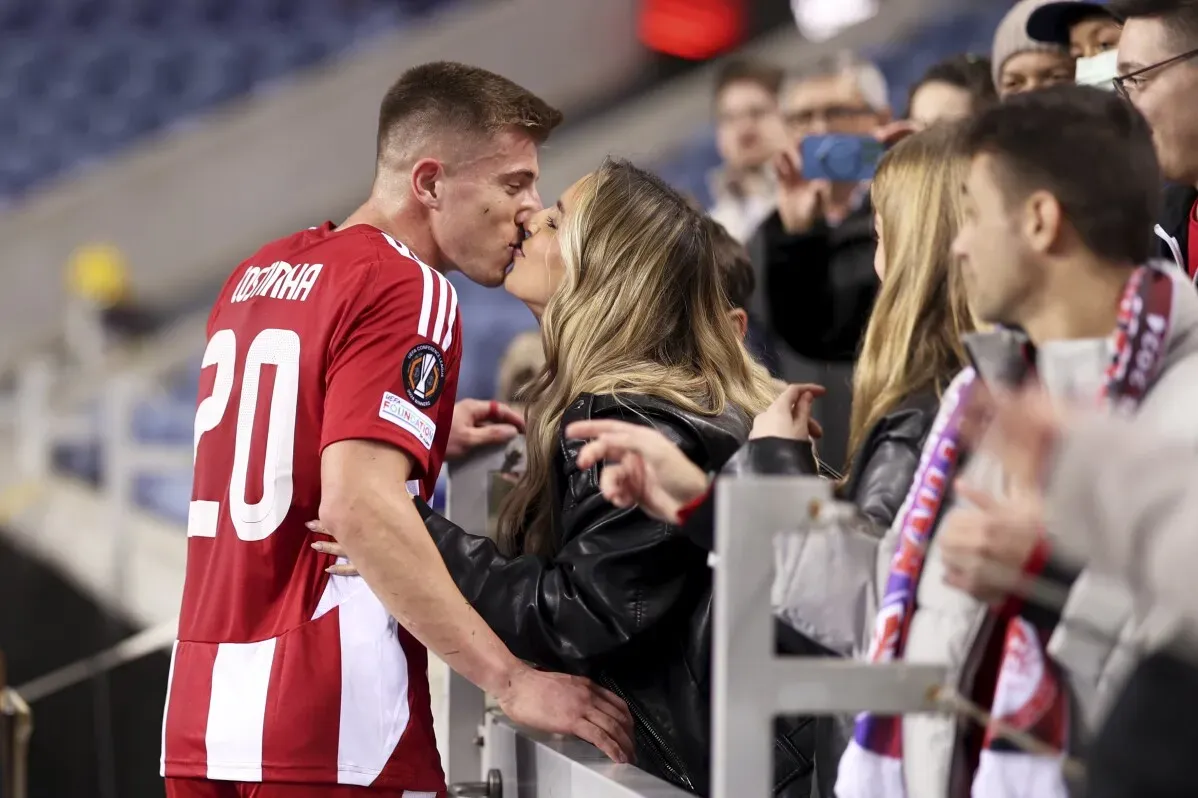 لاعب كرة قدم يحمل الرقم 20 يقبّل شريكة بعد المباراة، بينما الجماهير تشجع في مدرجات الملعب. تبرز سعادة اللحظة بعد الفوز في الدوري الأوروبي.
