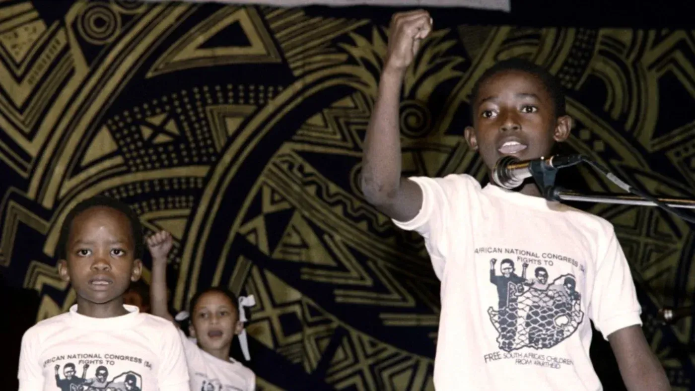 أطفال يتحدثون في حدث عام، يرتدون قمصان تحمل شعار "المؤتمر الوطني الأفريقي"، يعبرون عن مقاومة الظلم والمعاناة.