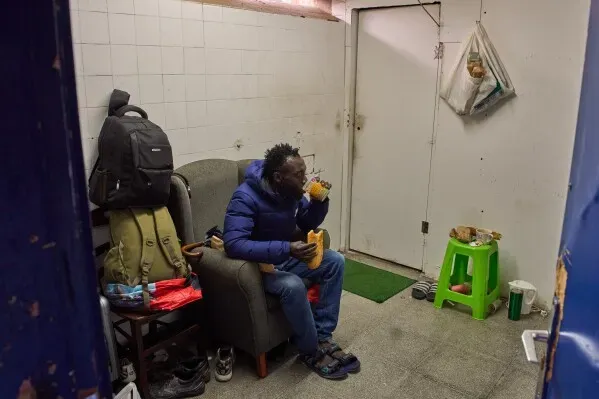 رجل يجلس في غرفة صغيرة، يتناول الطعام بينما تحيط به حقائب وأغراض شخصية، مما يعكس ظروف المهاجرين في بادالونا.