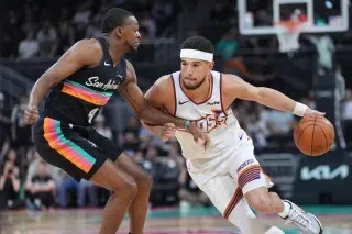 ديفين بوكر لاعب فينيكس صنز يتجاوز مدافع سان أنطونيو سبيرز خلال مباراة كرة سلة، مع التركيز على حركة اللعب والتنافس.