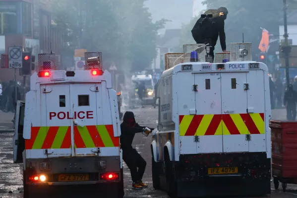 تصاعد الدخان في بلفاست خلال أعمال شغب، حيث يظهر رجال يرتدون أقنعة بالقرب من مركبات الشرطة وسط توتر اجتماعي متزايد.