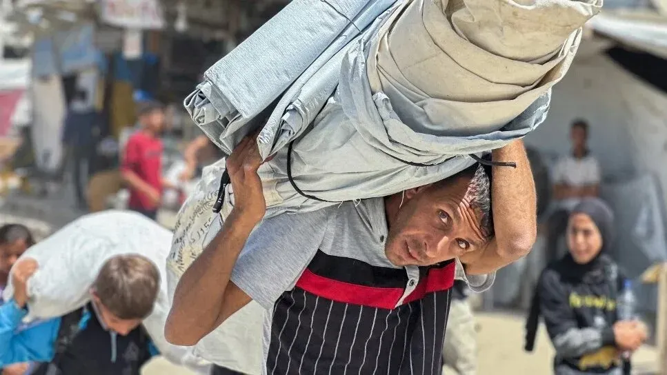 رجل يحمل أغطية ثقيلة فوق كتفه في منطقة خان يونس، محاط بأطفال ومشاهد من الفوضى، معبرة عن معاناة السكان في ظل الأوضاع الراهنة.